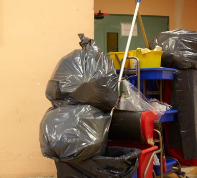 In the foreground, several large black rubbish bags, made of thick plastic with a slightly glossy finish, are stacked against a beige wall, occupying most of the image space. The bags are filled to capacity, causing them to bulge and appear tightly sealed at the top. To the right, a small red trolley or cart with a handle is visible, supporting additional waste containers, including a yellow plastic bin with a lid and a blue tray or container on top. Behind these, a dark grey or black trash bag leans against the wall. The background features a green door with a metallic handle and a small window, possibly part of a warehouse or storage area, suggesting a commercial or industrial environment in Leyton. The lighting is even and natural, highlighting the textures of the plastic bags and the contrast between the waste materials and the surrounding environment. The scene reflects a typical rubbish collection or waste removal setup, consistent with services provided by Business Waste Removal Leyton, in a setting such as a warehouse, back alley, or service area in Leyton, E10.