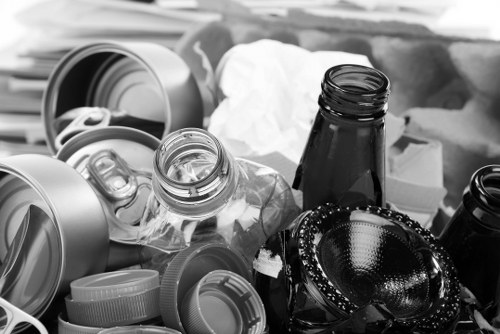 The image displays a collection of discarded waste items including multiple empty glass bottles with black caps, various plastic containers and lids of different sizes and shapes, some crumpled white paper, and a few scattered bottle caps. These objects are situated on a surface that appears to be an outdoor driveway or pavement, with a background that is blurred but suggests an urban or residential area. The waste items are haphazardly piled, indicating collection or removal in progress. The presence of glass and plastic materials reflects typical household or business rubbish, consistent with the services provided by Business Waste Removal Leyton in the Leyton area, within the local postcode region. Overall, the scene depicts typical waste that would be handled by professional rubbish removal firms specializing in clearing general household or commercial refuse in Leyton or nearby areas.
