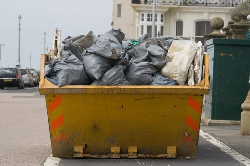 A large yellow industrial skip filled with black and grey rubbish bags, some of which are partially torn or crumpled, situated on a paved street next to a pavement in a residential or commercial area. The skip is positioned on the edge of the road, with a portion of an orange reflective safety stripe visible on its side. Surrounding the skip are parked vehicles and buildings with white facades and windows, indicating a typical urban setting, possibly in Leyton or nearby areas. The contents of the skip suggest waste removal services, like those offered by Business Waste Removal Leyton, are active in the area. The environmental lighting is natural, and the scene portrays a busy or routine rubbish collection site with community street elements in the background.