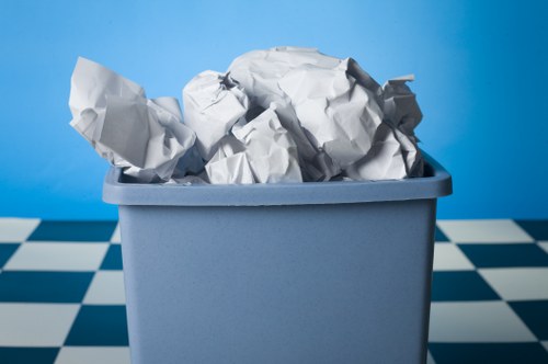 A light grey plastic bin filled with crumpled white paper and discarded sheets, positioned on a checkered floor with black and white tiles. The bin is set against a bright blue background, suggesting an indoor environment such as a kitchen or office space. The crumpled paper shows various folds and textures, indicating it is used waste material. The bin's surface appears smooth and matte, with a rectangular shape and rounded edges. The paper extends slightly above the rim of the bin, with some pieces leaning to one side. The scene appears clean and organized, typical of waste removal for small-scale rubbish collection services in Leyton or nearby areas. Business Waste Removal Leyton often manages such waste types, which might include office or domestic paper waste, collected from urban residential or commercial properties in the local postcode area.