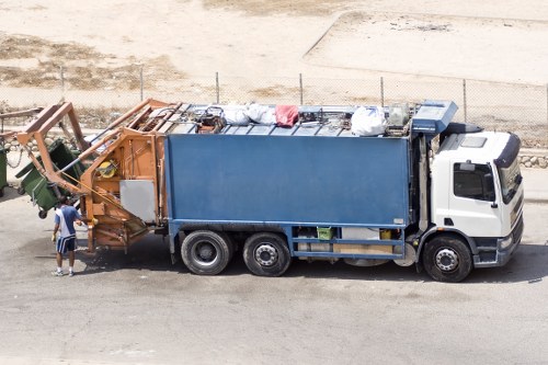 A large waste collection truck from Business Waste Removal Leyton is parked on a paved surface, with its blue waste containment body clearly visible and filled with various waste materials, including bags and loose items. The front cab of the truck is white, with a dark windshield and side mirrors, and positioned on the right side of the image. In the foreground, a worker wearing a high-visibility vest and jacket stands beside a green mechanical arm attached to the front of the truck, which appears to be used for loading or unloading waste. The background shows a gravel or dirt area with minimal vegetation, a low fence, and an uneven ground surface, suggestive of an open industrial or waste management yard. The scene is lit naturally, with bright, clear lighting indicating daytime, and the environment indicates an outdoor rubbish removal operation typical of Leyton or nearby London suburbs, managed by Business Waste Removal Leyton.
