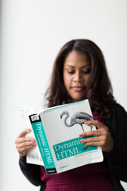 A woman with medium-length dark hair and light brown skin is standing indoors against a plain white wall, holding a book titled 'Dynamic HTML' by O'Reilly. She is wearing a dark blazer over a burgundy