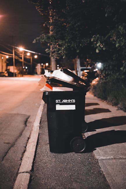 Two large black plastic bags of household waste, placed on the edge of a paved street, are visible in front of a tall, dark wooden fence with vertical slats. The bags appear to be filled with miscella