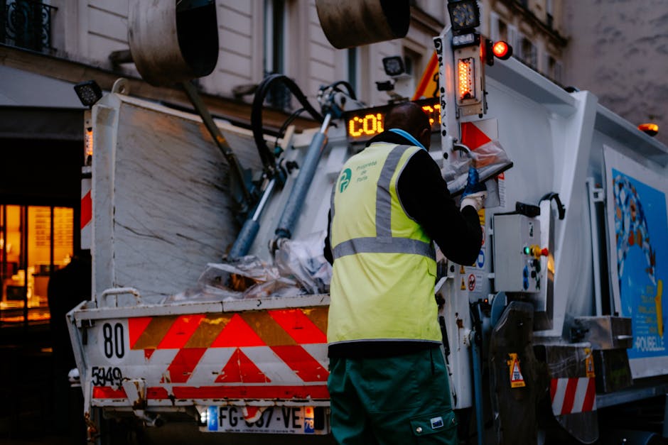 A waste collection vehicle parked on a city street during twilight, with its rear hopper open filled with mixed waste materials such as paper, plastic, and household refuse. The vehicle features red a